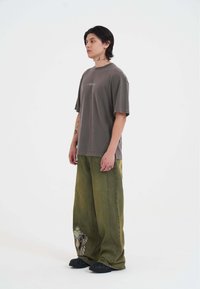 Grå oversized t-shirt med slidte kanter, parret med løse grønne bukser med løve-grafik. Sorte fodtøj; tatoveringer synlige på armene.