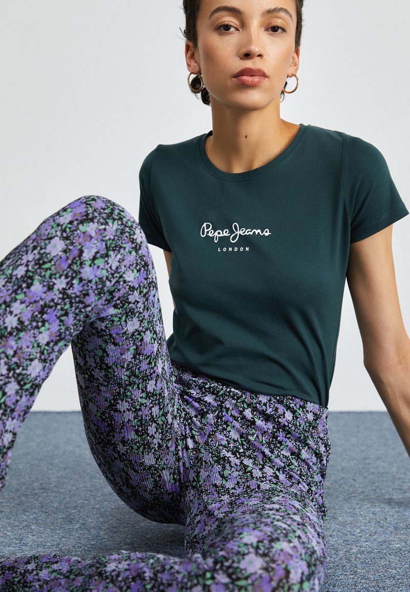 Tamsiai žalia marškinėliai su balta "Pepe Jeans London" logotipu; derinami su margais gėlių tipo leggingais purpurine ir žalia spalvomis ant tekstūruoto paviršiaus.