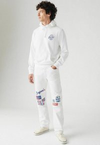 Witte katoenen hoodie met een logo, gecombineerd met witte broeken voorzien van kleurrijke tekstgraphics. De schoenen zijn lichtbeige sneakers.