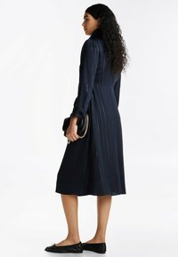 Vestido azul marino con rayas verticales, mangas largas y cintura ajustada. La tela parece suave con una silueta fluida. Combinado con zapatos planos negros.