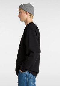 Felpa nera con scollatura a girocollo, maniche lunghe e polsini a costine, abbinata a jeans blu. Viene indossato un berretto di maglia grigia.