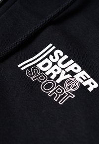 Tejido de sudadera negra con capucha que presenta un logo blanco con el texto "SUPERDRY" y "SPORT". Incluye tres franjas paralelas por encima del logo.
