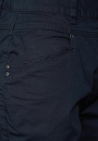 Jean bleu foncé avec une texture lisse, comportant deux boutons métalliques et des détails de couture le long des poches et des coutures.