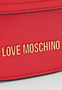 Červená koženka s zlatým nápisem "LOVE MOSCHINO", s hladkým povrchem a kontrastním černým prošíváním podél okraje.