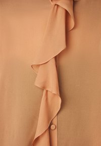 Peachfarget bluse med volangkrage og knappestolpe. Laget av glatt stoff med en flytende tekstur og subtil glans.