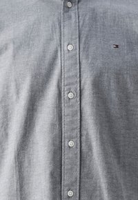 Grå skjorta med knappar, gjord av lätt tyg med en slät textur, sex knappar och en liten broderad logotyp på bröstet.