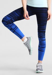 Blå mönstrade tights med mörka ränder, tillverkade av elastiskt material, ihop med grå träningsskor med gröna accenter.