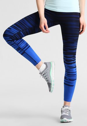 Niebieskie legginsy w wzory z ciemnymi paskami, wykonane z elastycznego materiału, zestawione z szarymi butami sportowymi z zielonymi akcentami.