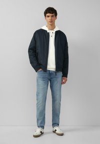 Marine bomberjack met witte capuchon, lichtblauwe jeans en schoenen met donkere accenten. Gladde stof, ontspannen pasvorm, ritssluiting en zakken.