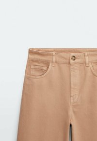 Pantalon en denim beige taille haute avec bouton frontal, braguette zippée, passants pour ceinture et poches avant visibles sur fond blanc.