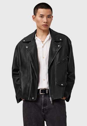 Veste de moto en cuir noir avec quincaillerie argentée, poches zippées et un large col. Associée à une chemise blanche et un jean foncé.