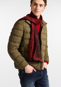 Olivengrøn pufferjakke med quiltet tekstur, r�ød tørklæde med sorte mønstre og tætsiddende blå jeans.