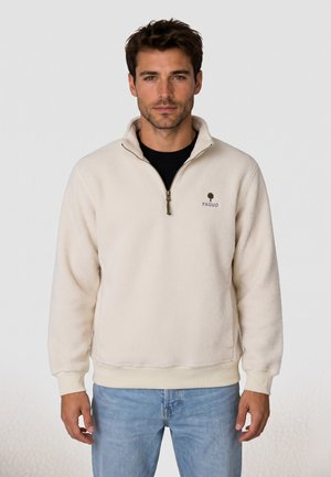 Hombre con un jersey polar crema con media cremallera y logo de "Faguo" y pantalones vaqueros azul claro, de pie frente a un fondo liso.