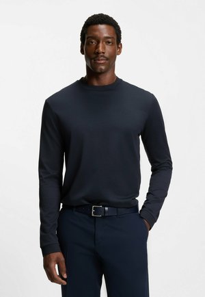 Ein Mann trägt ein eng anliegendes, langärmeliges, dunkelblaues Hemd, das in passende dunkelblaue Hose mit einem schwarzen Gürtel gesteckt ist, und steht vor einem schlichten Hintergrund.