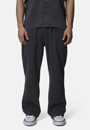 Uomo con pantaloni larghi di tessuto scuro e camicia abbinata, indossando sneakers bianche, in piedi davanti a uno sfondo chiaro e uniforme.