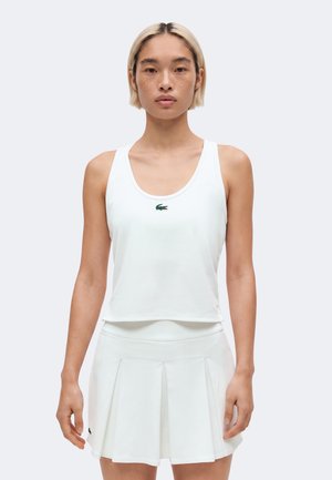 Femme aux cheveux blonds courts portant un haut sans manches blanc et une jupe plissée blanche avec un logo crocodile vert sur la poitrine et la jupe.