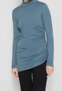 Langärmliges Oberteil aus weichem blauen Stoff mit hohem Kragen und gerafften Seitendetails, ausgestattet mit asymmetrischem Saum und schlankem Design.