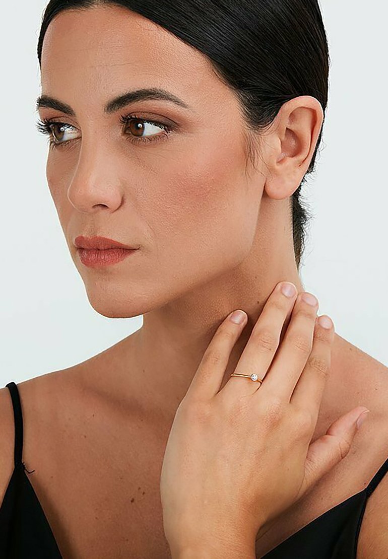 Goldring mit einem kleinen klaren Edelstein, der an einer Hand getragen wird. Das Haar des Modells ist nach hinten gebunden, sie trägt ein schwarzes Oberteil und einen hellen Hintergrund.