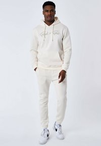 Completo composto da hoodie e pantaloni jogger color crema in tessuto morbido, con tasca frontale e dettaglio con cordino; abbinato a sneakers bianche.