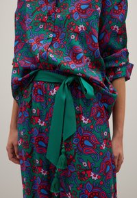 Kleurige bloempatronen blouse met groene achtergrond, voorzien van rode en blauwe accenten, afgewerkt met een bijpassende groene knoopceintuur en kwastdetail.