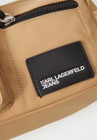 Tan nylon bag featuring a black leather patch with embossed "KARL LAGERFELD JEANS" logo. Includes a zippered pocket and neutral stitching.