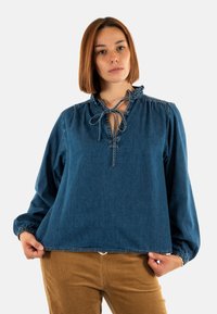 Blusa in denim di colore blu intenso con scollatura a volant, dettaglio con fiocco e maniche lunghe a sbuffo. Morbido al tatto e con una vestibilità comoda.