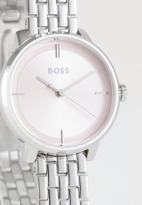 Montre en métal argenté avec une finition polie, dotée d'un cadran rose pâle, de fines aiguilles et d'un bracelet texturé avec un design tissé.