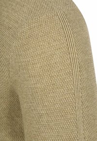 Texturierter beiger Strickstoff mit einer detaillierten Rippennaht, die vertikal im Schulterbereich eines Kleidungsstücks verläuft.
