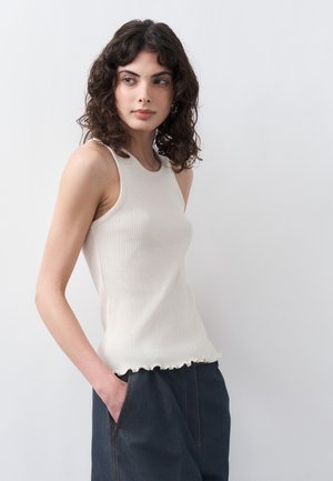 Femme aux cheveux bouclés et foncés, portant un haut blanc sans manches à côtes et un pantalon foncé, debout avec une main dans la poche, regardant sur le côté.