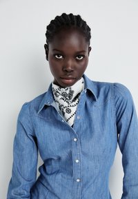 Niebieska koszula dżinsowa, jasna, z zapięciem na guziki i spiczastym kołnierzykiem. Posiada teksturowaną tkaninę oraz białą chustę w czarny kwiatowy wzór.