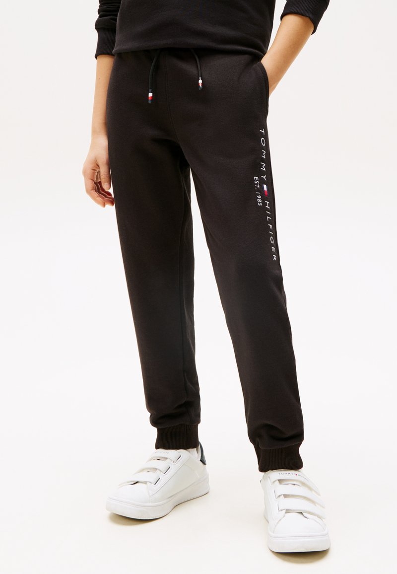 Pantalons de survêtement noirs en tissu doux, avec une taille élastique à cordon de serrage, des poches latérales et une marque en texte blanc le long de la jambe.
