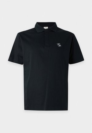 Άνδρας στέκεται φορώντας μαύρο polo μπλουζάκι με κοντά μανίκια, ελιάς πράσινα cargo παντελόνια και μαύρα-άσπρα loafers σε απλό γκρι φόντο.