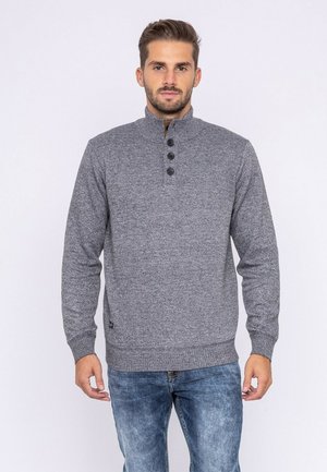 Homme portant un pull gris en maille avec un col montant et quatre boutons noirs, associé à un jean bleu, debout devant un fond uni.