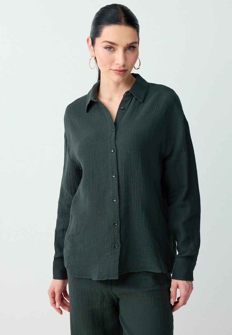 Femme aux cheveux foncés portant des boucles d'oreilles créoles dorées, une chemise vert foncé à manches longues boutonnée ainsi qu'un pantalon assorti sur un fond clair.
