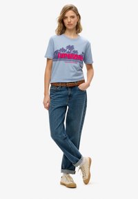 Jasnoniebieska koszula T-shirt z różowym napisem "SUPERDRY", w połączeniu z jeansami z wysokim stanem w niebieskim kolorze oraz beżowymi sneakersami, z podwiniętymi rękawami i plecionym paskiem.