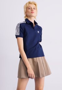 Polo-shirt navy avec des rayures blanches sur les manches, arborant un petit logo. Porté avec une jupe plissée beige, mettant l'accent sur des lignes épurées et une coupe moderne.