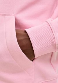 Vaaleanpunainen collegepaita resorihihoilla, jossa on etutasku. Lähikuva näyttää tasaisen pinnan ja käden taskussa.