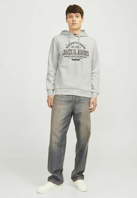 Grauer Hoodie mit einem Frontdruck, Kordelzugkapuze und gerippten Bündchen. Kombiniert mit lockeren blauen Jeans und weißen Turnschuhen.