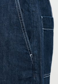 Σκούρο μπλε τζιν ύφασμα με λευκές ραφές, που δείχνει μια εμπρόσθια τσέπη και μια πλευρική τσέπη με ενισχυμένες ραφές.