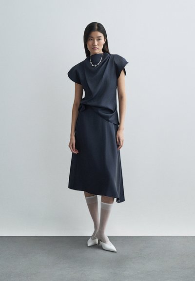 A navy blue dress with cap sleeves, knee-length, and a relaxed fit. Paired with white heeled shoes and knee-high sheer socks. Simple necklace.