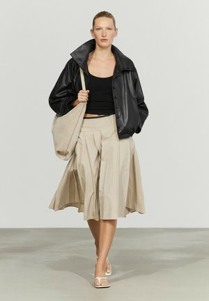 Donna che cammina indossando un top nero corto, giacca di pelle nera, gonna beige plissettata, sandali bianchi e porta una borsa beige a tracolla.