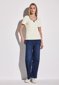 Top corto a maniche corte di colore giallo chiaro con coste e bordi neri, abbinato a jeans larghi blu e sneakers bianche, su uno sfondo semplice.