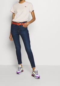 Ljusrosa bomullströja med liten röd logotyp, mörkblå tajta jeans, blank röd bälte och flerfärgade sneakers med lila accenter.