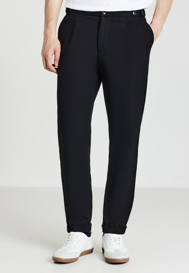Pantalon noir avec une coupe droite, comportant deux poches avant et une texture lisse. Associé à des baskets blanches sur un fond neutre.