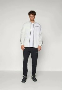 Hvit lett jakke med svarte detaljer og et merke, kombinert med svarte bukser og mørkt fottøy, designet for aktivt bruk.