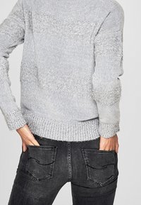 Hellgrauer Pullover mit flauschigen horizontalen Streifen, geripptem Saum und Bündchen. Kombiniert mit schwarzen Jeans, die auf der Rückseite Taschen mit geschwungenen Nähten haben.