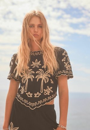 Svart kort topp med krämfärgad brodyr som har palmträd och blommönster, med vågiga kanter och korta ärmar, kombinerad med en svart underdel.