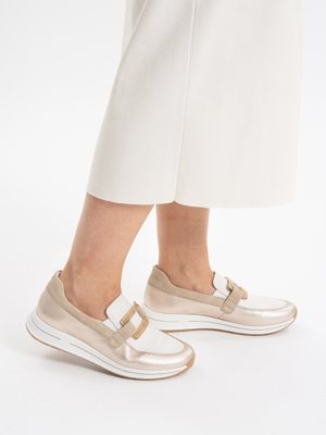 Beine in weißen, verkürzten Hosen und beige-weißen Slip-on-Sneakern mit dicken weißen Sohlen und dekorativen Riemen.