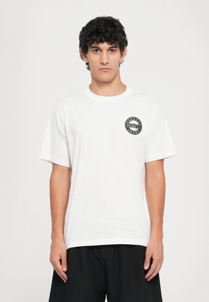 Bela bombažna majica s kratkimi rokavi in okroglim izrezom, s črnim krožnim logotipom na levem prsnem predelu, ki vsebuje besedilo "VERSACE COUTURE JEANS."