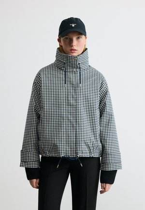 Žena v černobílé kostkované bundě s vysokým límcem, černých kalhotách, černé kšiltovce Barbour a černých bezprstých rukavicích, stojící před jednobarevným pozadím.